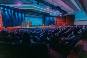 Equipe reunida em convenção corporativa no WTC Events Center, com palco iluminado, projeções e ambiente moderno.