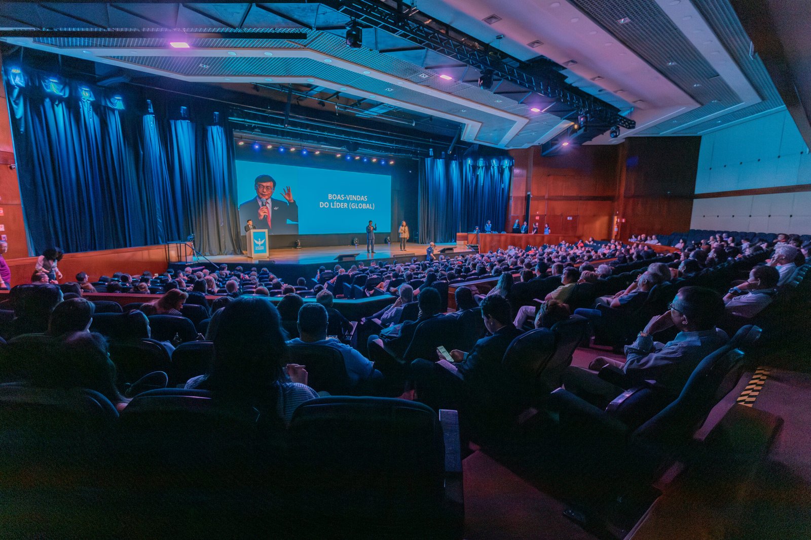 Equipe reunida em convenção corporativa no WTC Events Center, com palco iluminado, projeções e ambiente moderno.